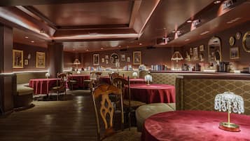 Un restaurante tenuemente iluminado con manteles rojos, sillas de madera y una zona de bar con libros y objetos decorativos.