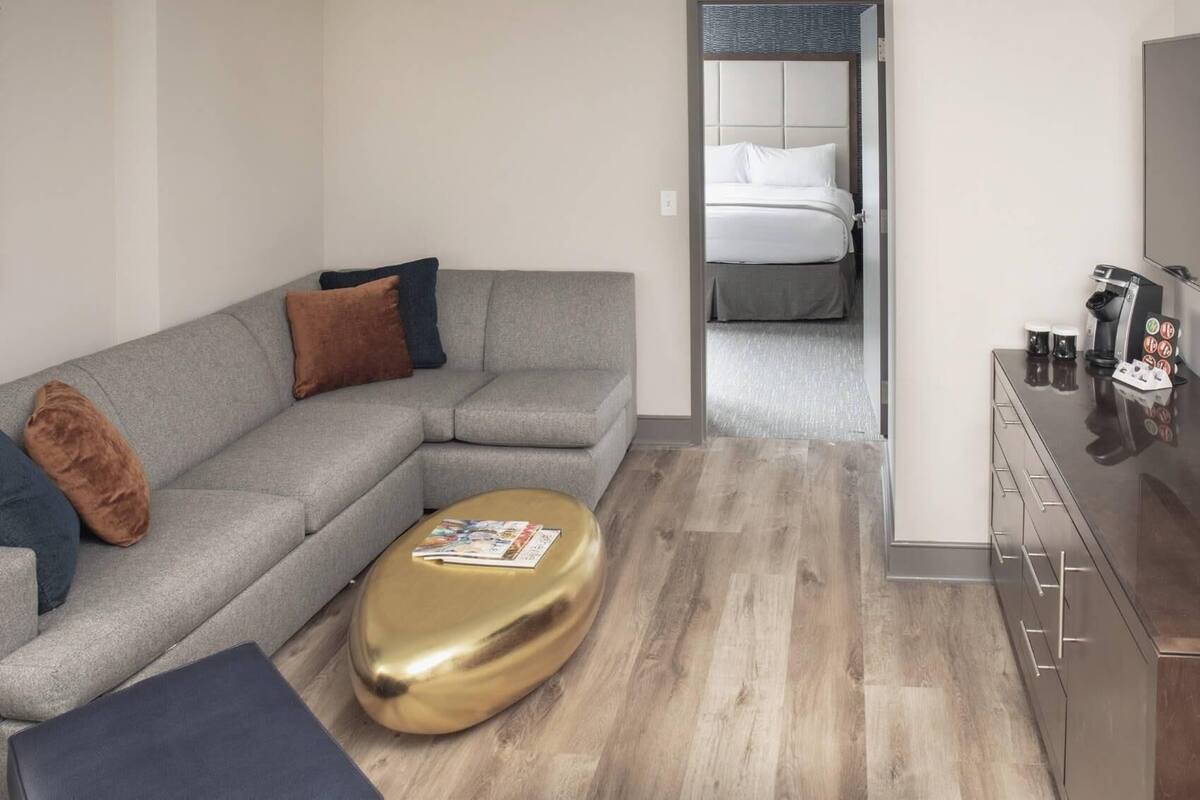 Une chambre d’hôtel moderne avec un canapé gris, un pouf doré, un plancher en bois, un lit avec une literie blanche et un minibar.