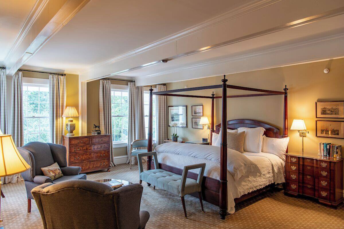 A four-poster bed in a well-lit bedroom with a large window, a wooden dresser, and a chair.