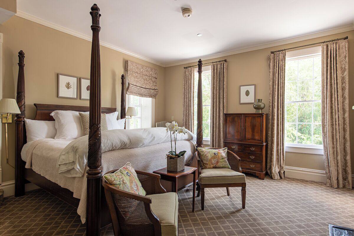 A four-poster bed with a nightstand, a chair, and a wooden cabinet in a room with large windows and a checkered floor.