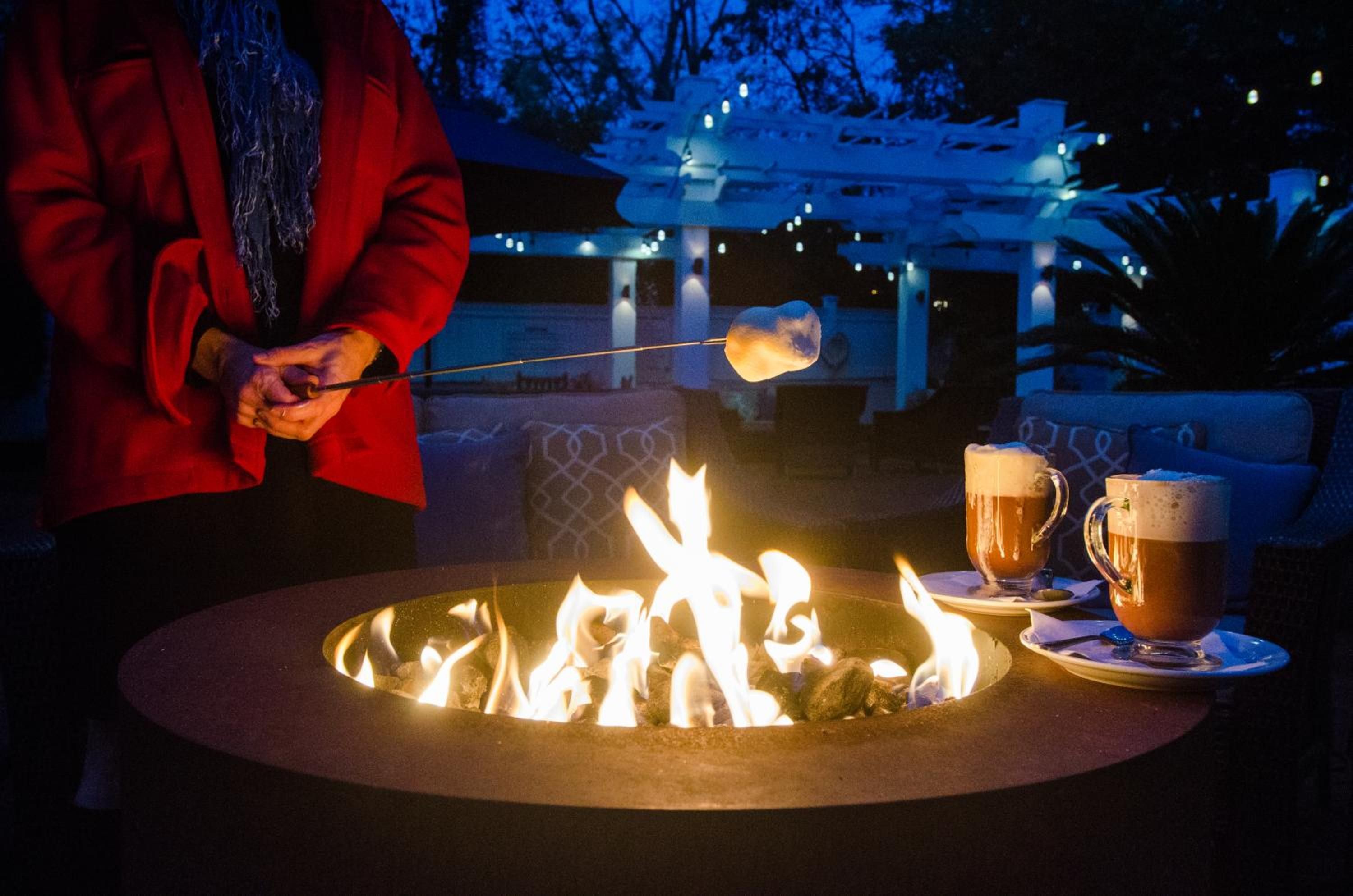 A person roasting marshmallows over a fire pit with drinks on the side.