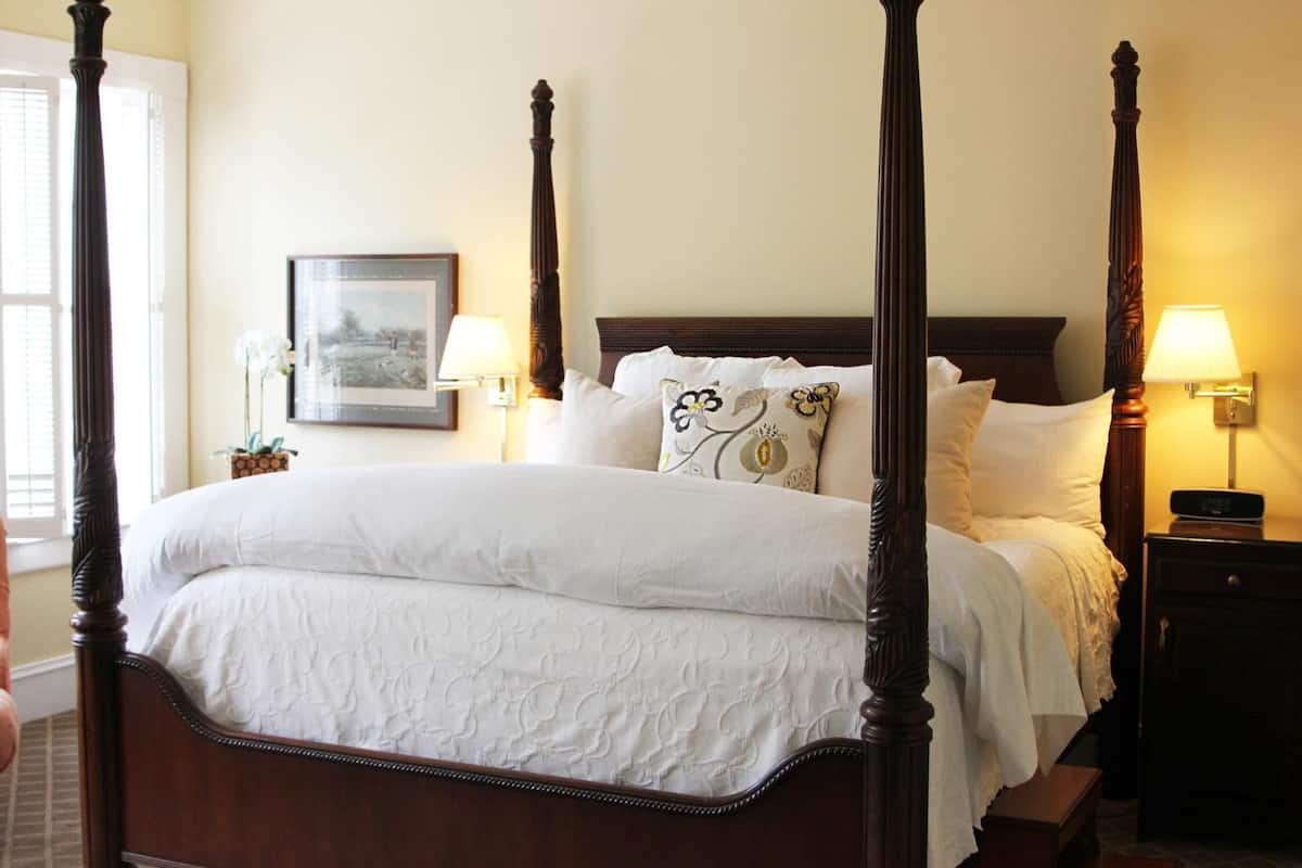 A four-poster bed with white linens, a framed picture on the wall, and two bedside lamps.