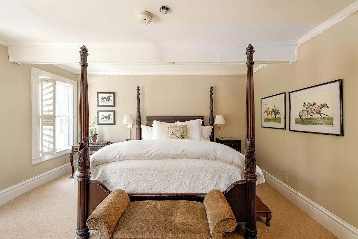 Een hemelbed met houten frame en witte beddengoed, een nachtkastje met een lamp, en twee ingelijste foto's aan de muur.