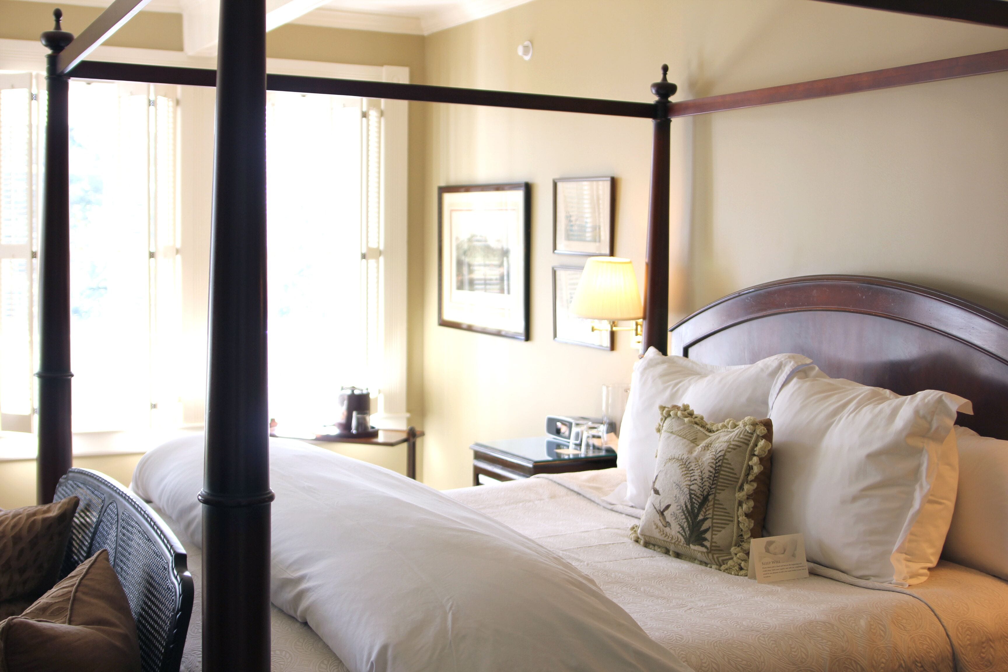 A four-poster bed with white linens, a wooden headboard, and a nightstand with a lamp.