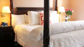 A four-poster bed with white linens, a wooden headboard, and a bedside table with a lamp and a vase of flowers.
