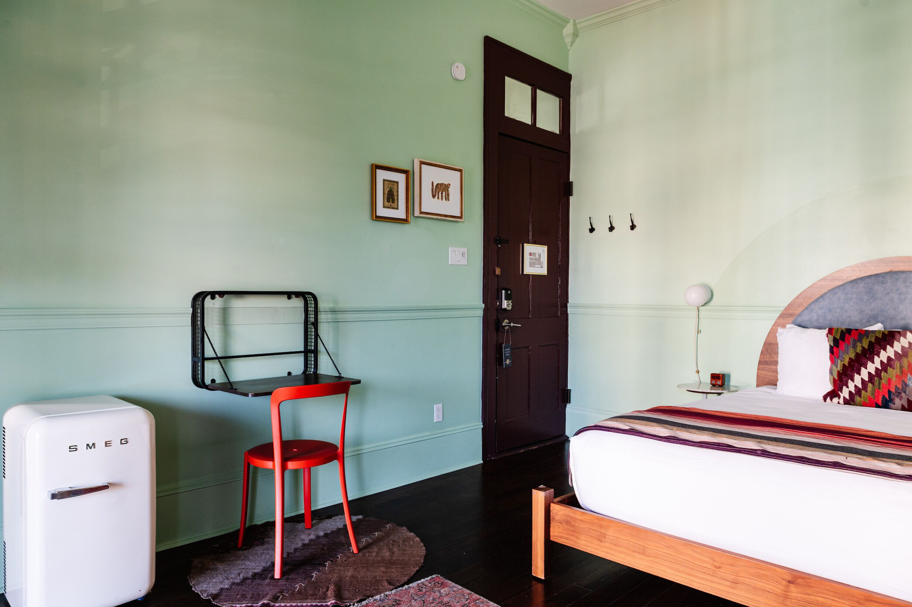 A bedroom with a bed, a red chair, a white fridge, a door, and two framed pictures on the wall.