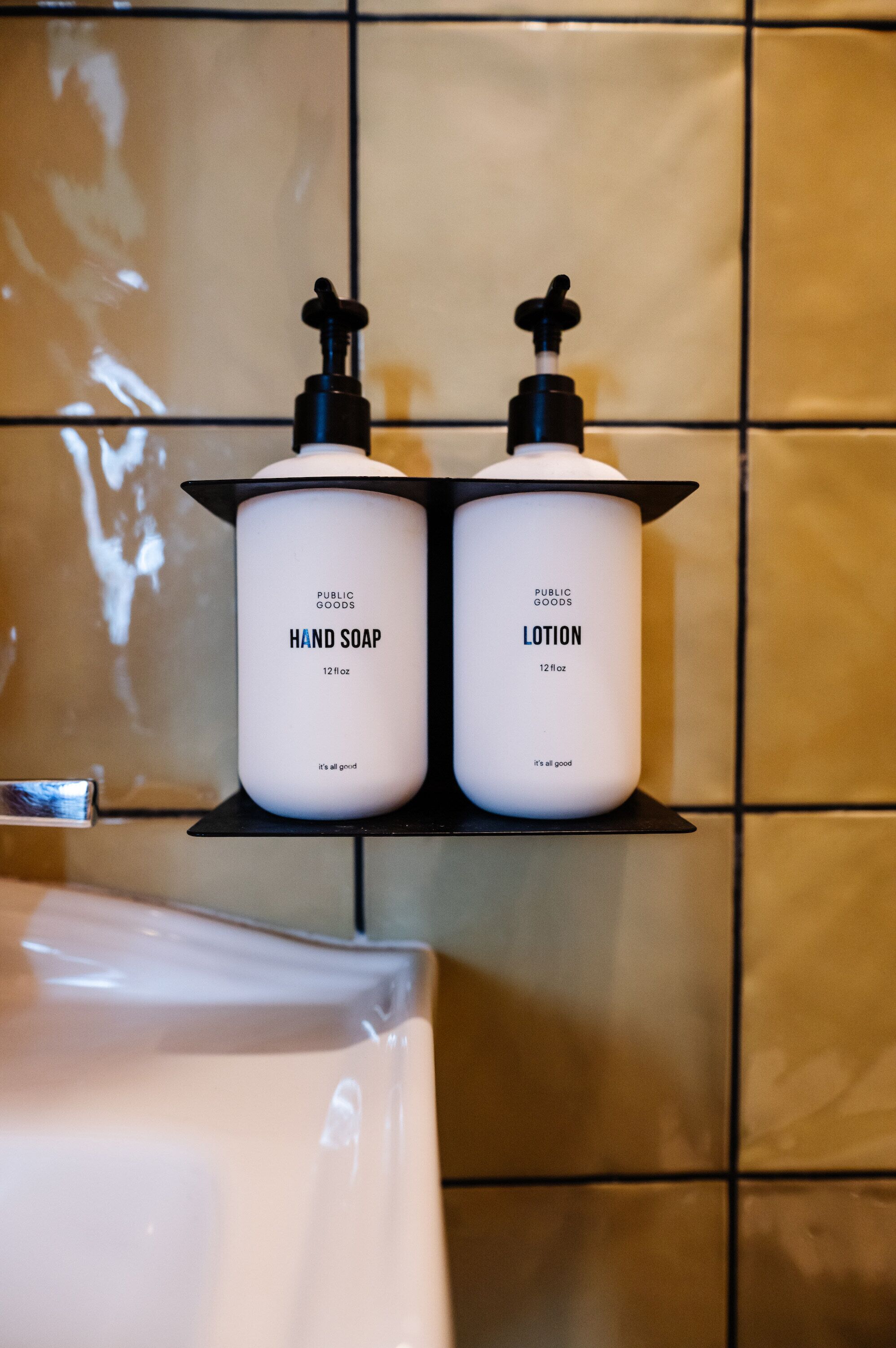 Deux récipients blancs portant les mentions « SAVON POUR LES MAINS » et « LOTION » de la marque « PUBLIC GOODS », placés sur une étagère noire dans une salle de bain aux murs carrelés.