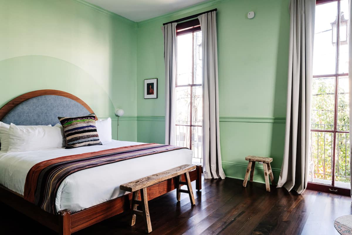 A room with a large bed, a wooden bench, a small stool, and a window with curtains.