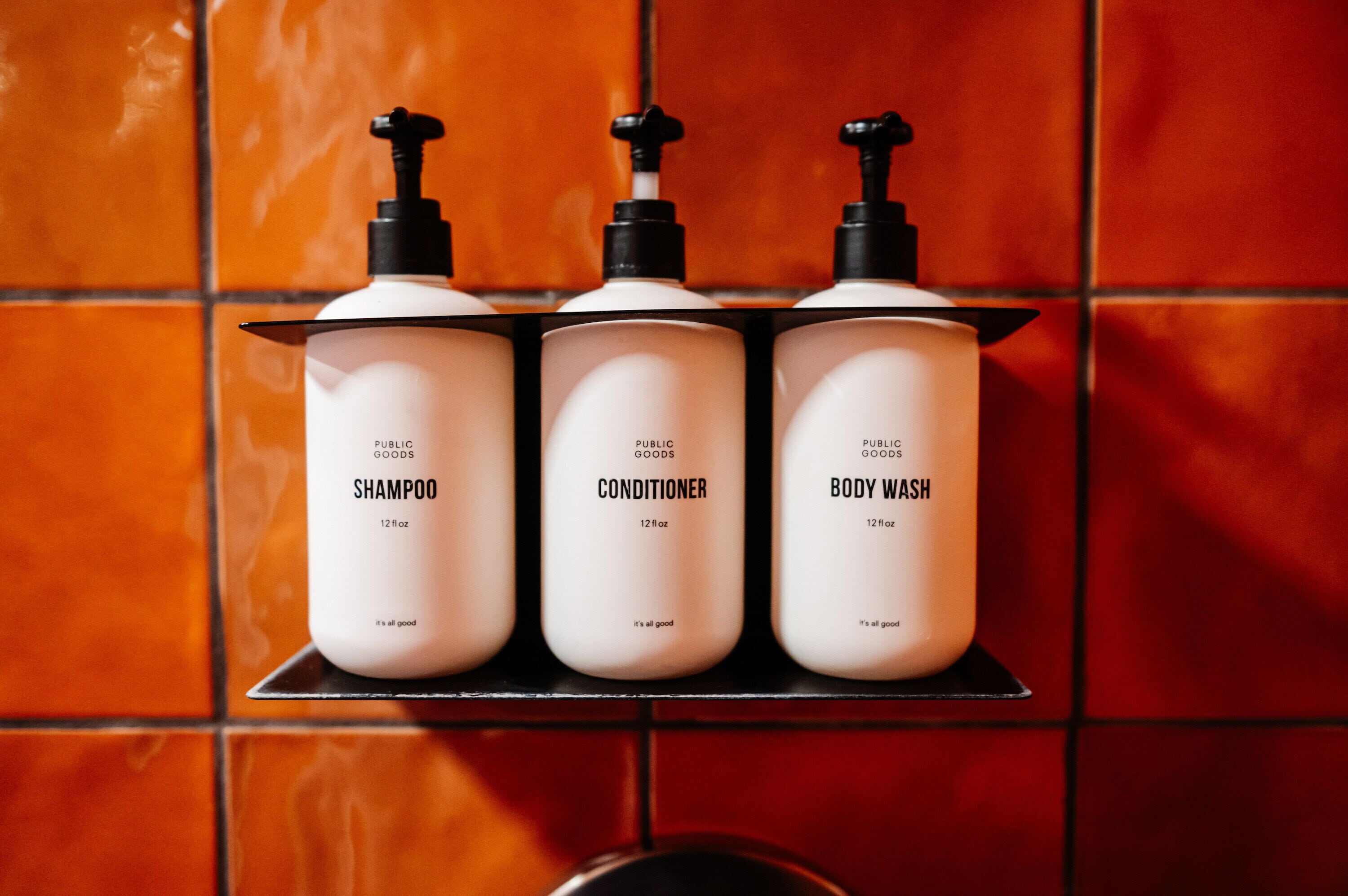 Three bottles of Public Goods products on a shelf against a tiled wall.