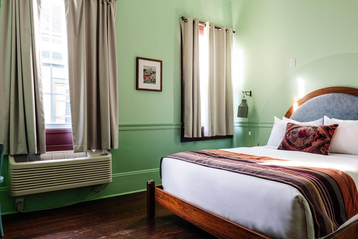 A room with a bed, a window with curtains, a picture on the wall, and a wall-mounted lamp.
