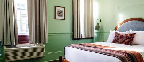 A room with a bed, a window with curtains, a picture on the wall, and a wall-mounted lamp.