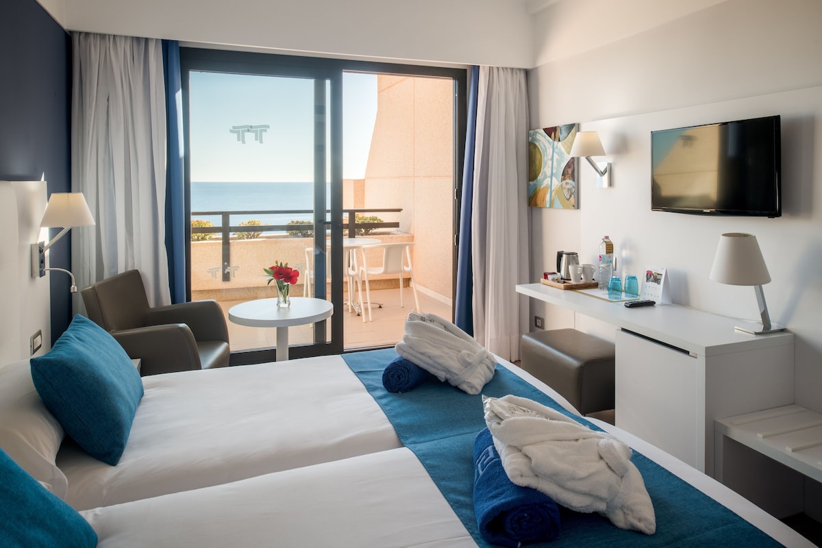 A hotel room with a bed, a desk, a TV, and a balcony with a view of the sea.