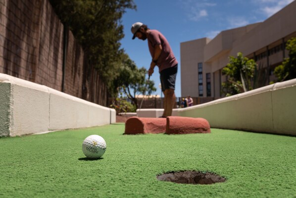 A golf putting green with a golf ball in the foreground, a sand trap, and a person in the background. - Alexandre Grand Teguise Playa (Teguise)