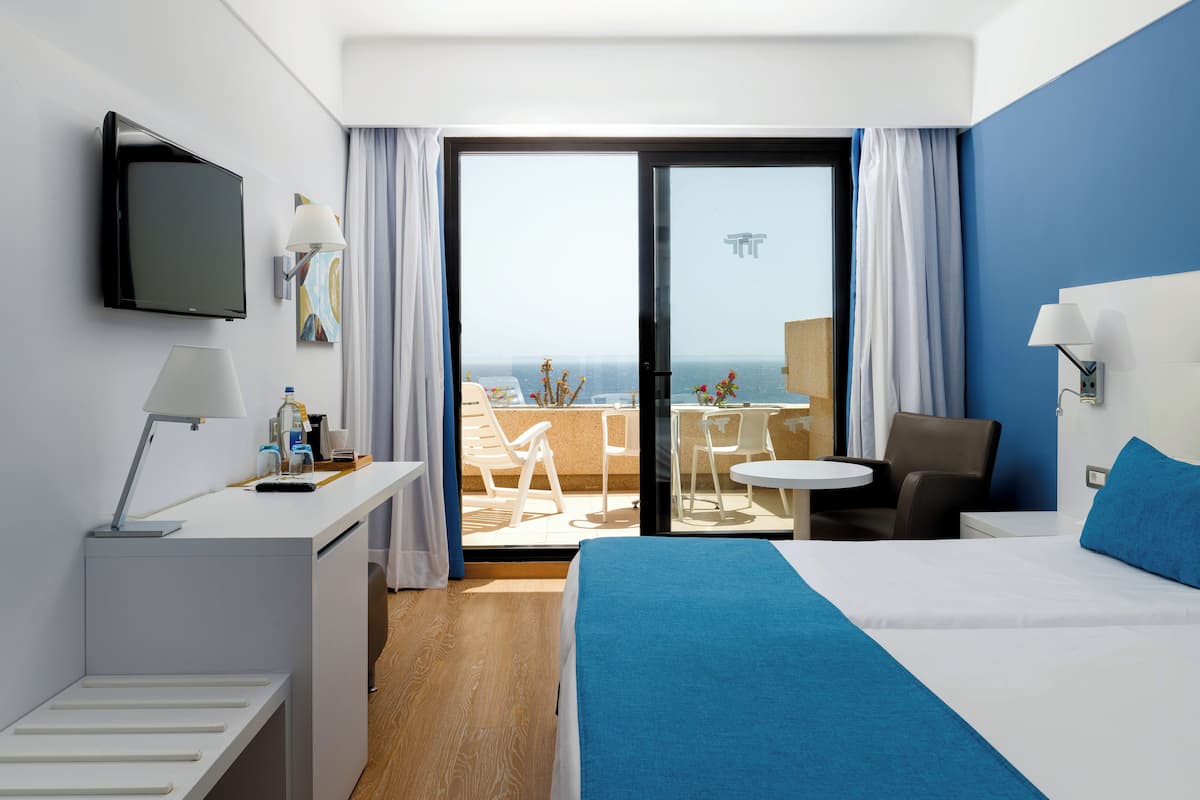 A hotel room with a bed, a desk, a TV, a balcony with chairs and a table, and a view of the sea.