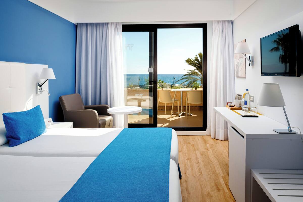 A hotel room with a bed, a desk, a chair, a small table, and a balcony with a view of the ocean.