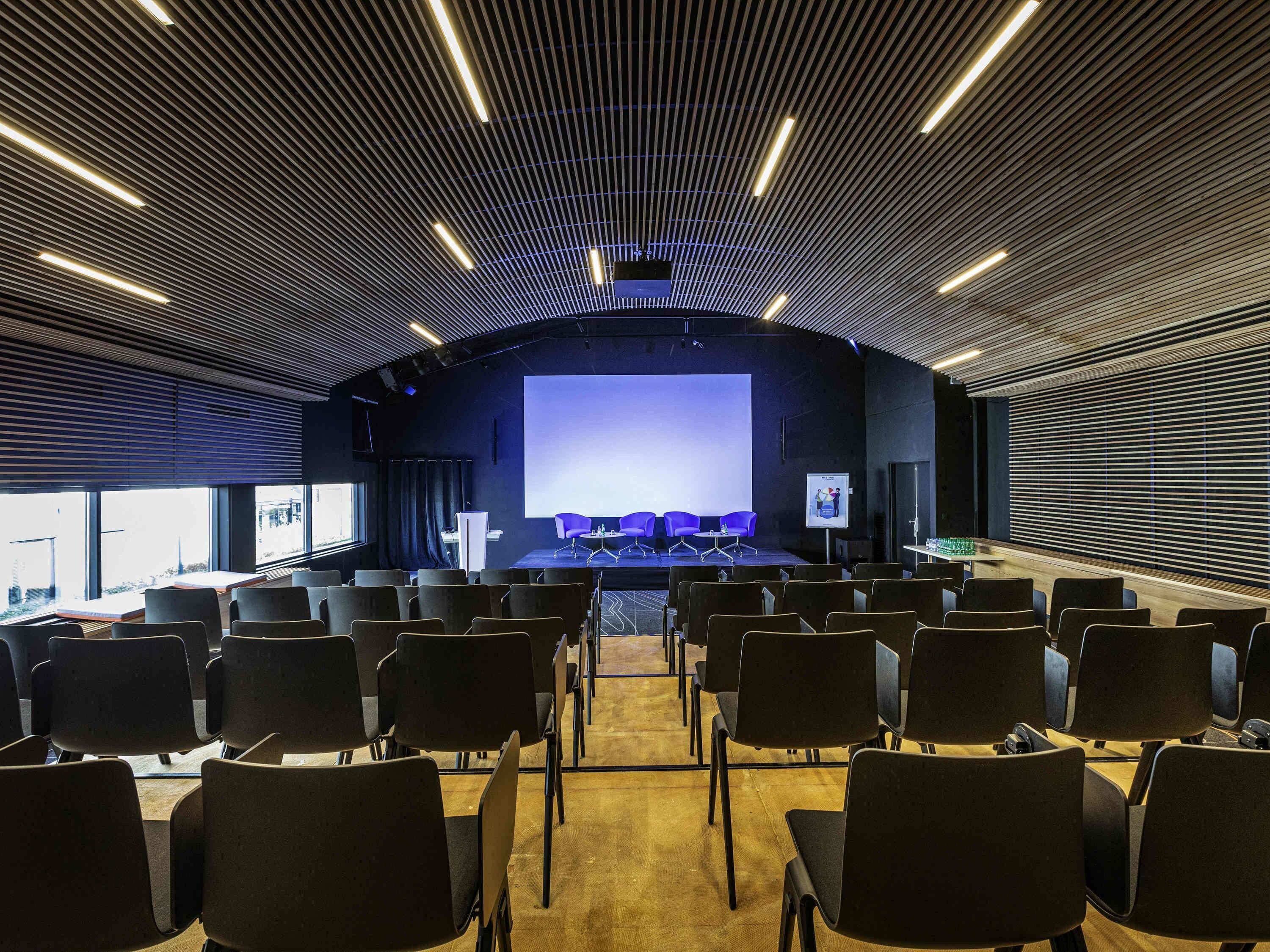 A conference room with a stage, rows of chairs, and a large screen.