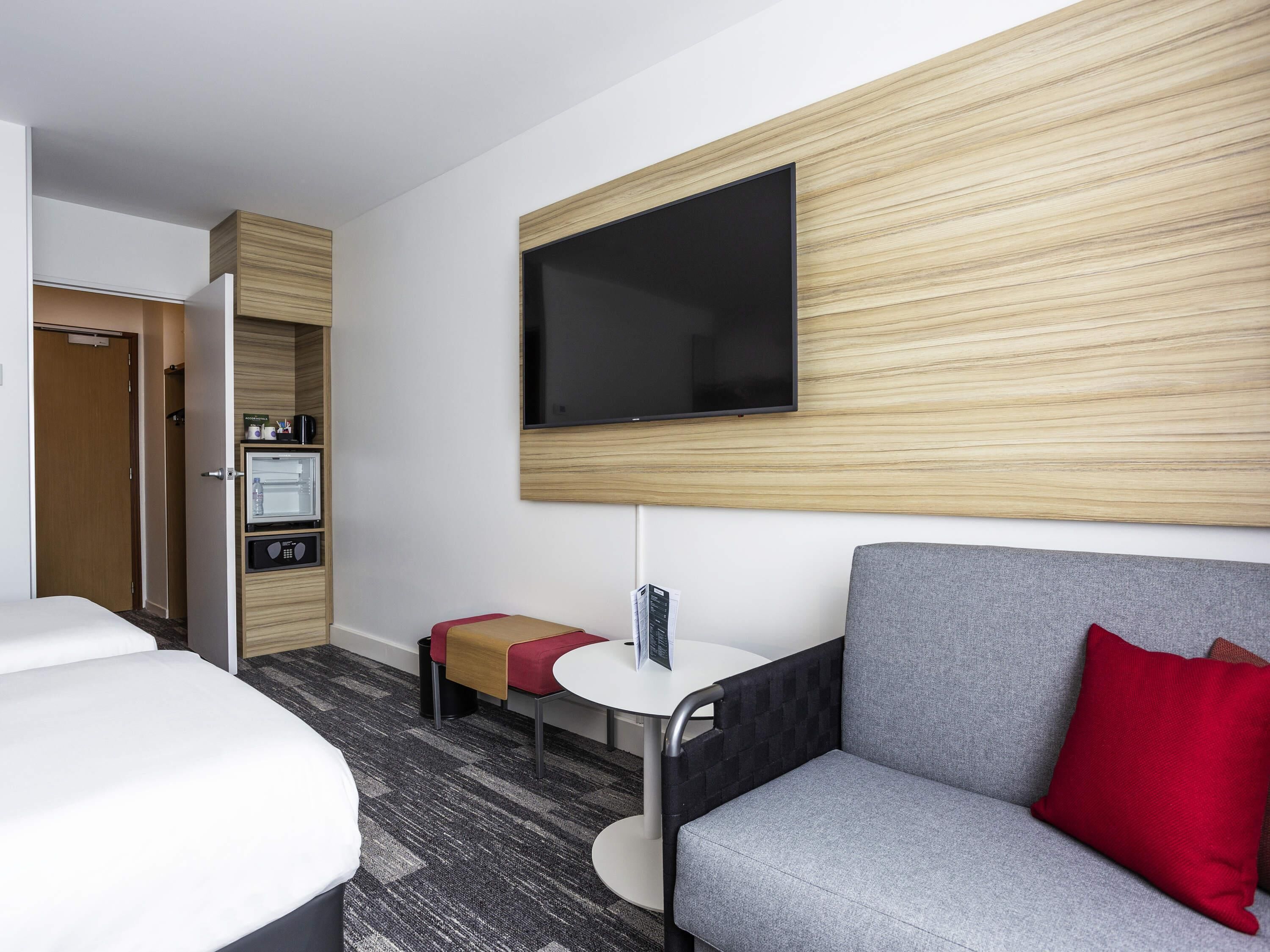 A hotel room with a bed, a sofa, a small table, a TV mounted on the wall, a minibar, and a door leading to a bathroom.
