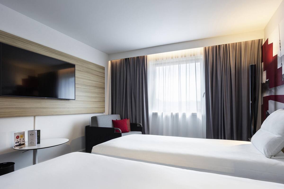A hotel room with two beds, a TV, a small table, and a window with curtains.