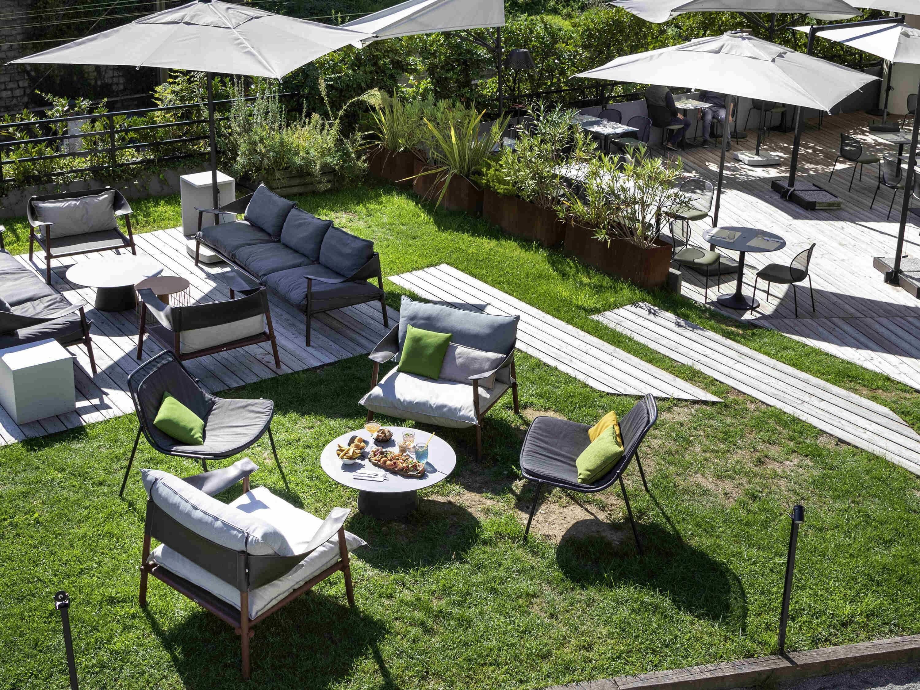 An outdoor patio with a sectional sofa, chairs, and tables, surrounded by greenery and umbrellas.
