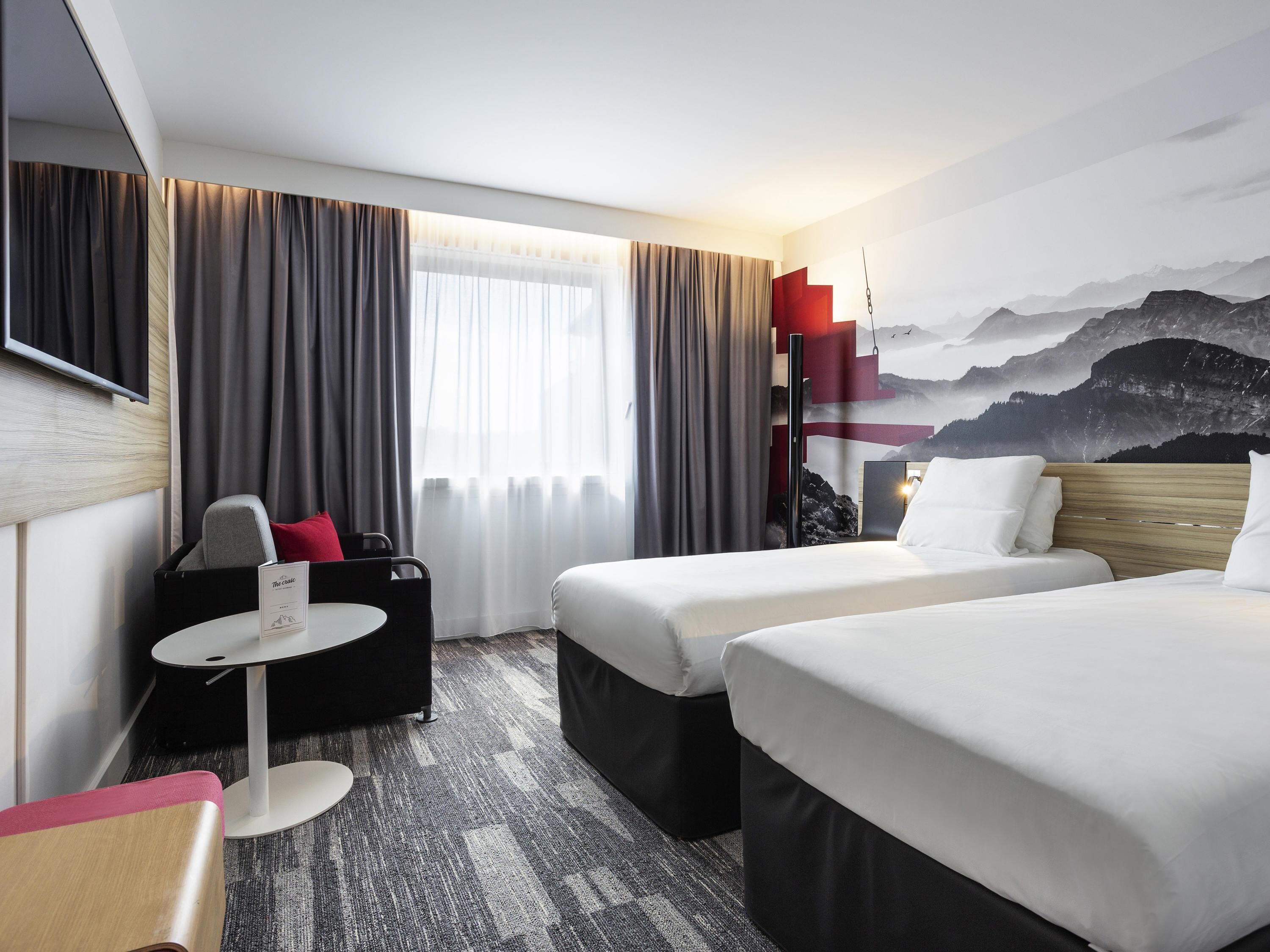 A hotel room with two beds, a desk, a chair, a small table, and a large window with curtains.