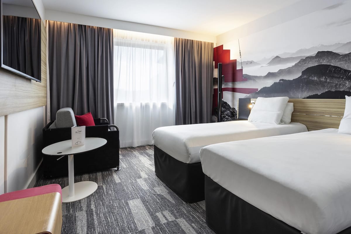 A hotel room with two beds, a desk, a chair, a small table, and a large window with curtains.