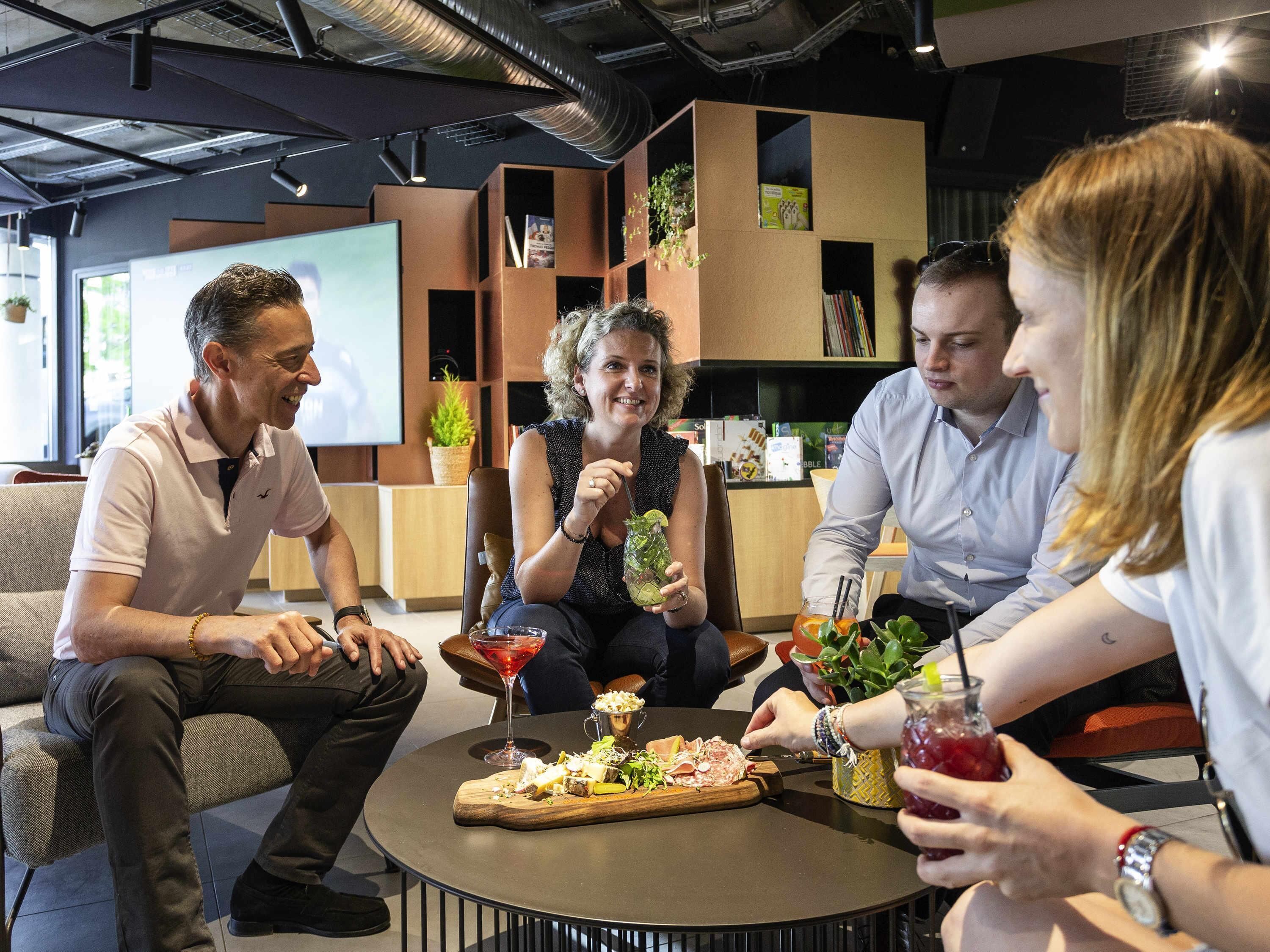 A group of people seated around a round table with a variety of food and drinks, in a modern indoor setting with bookshelves and a large screen in the background.
