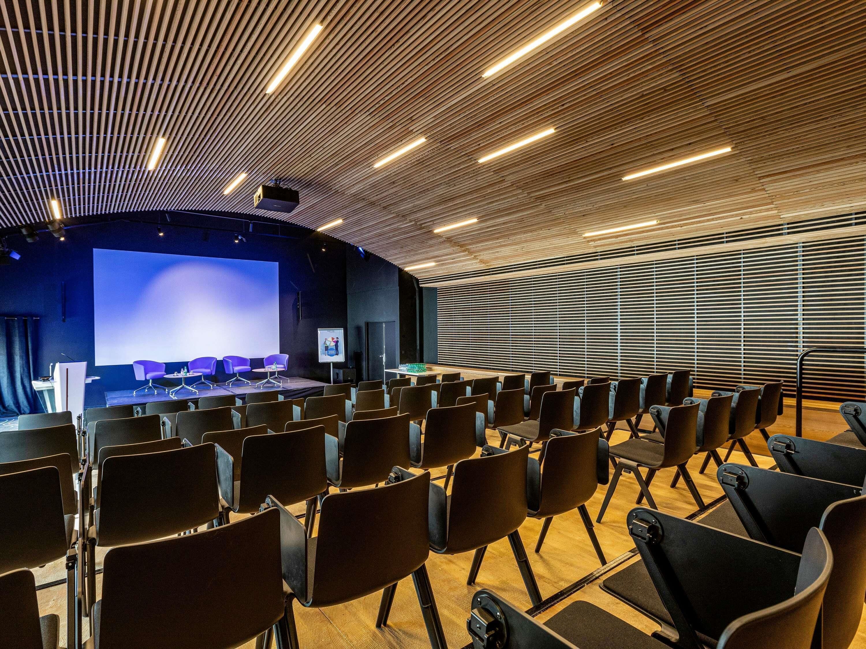A conference room with rows of chairs, a stage with a large screen, and a wooden slatted ceiling.