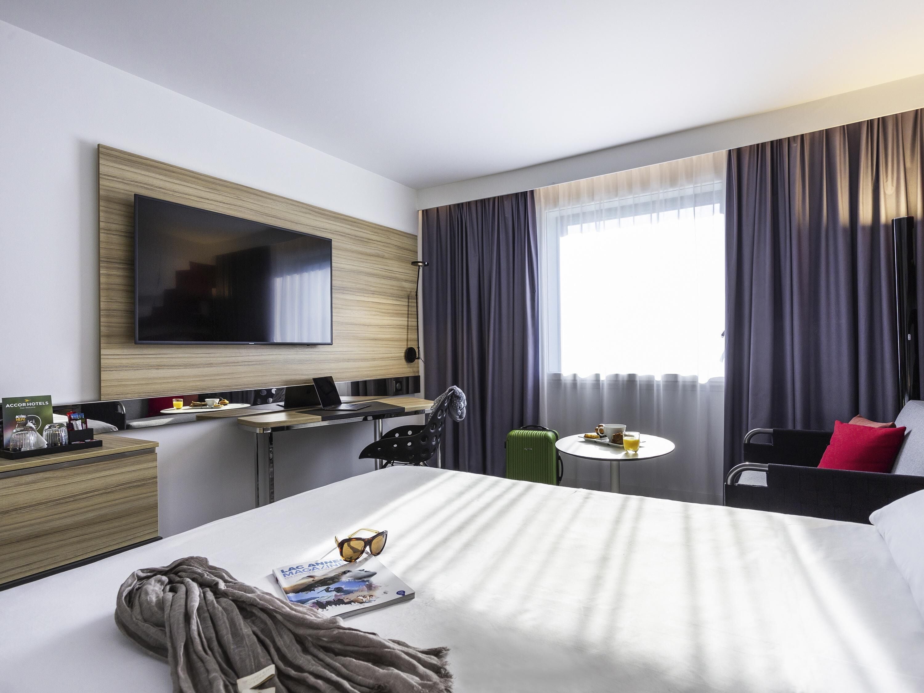 A hotel room with a bed, a desk with a laptop, a TV, a small table with a cup and a glass, and a suitcase.