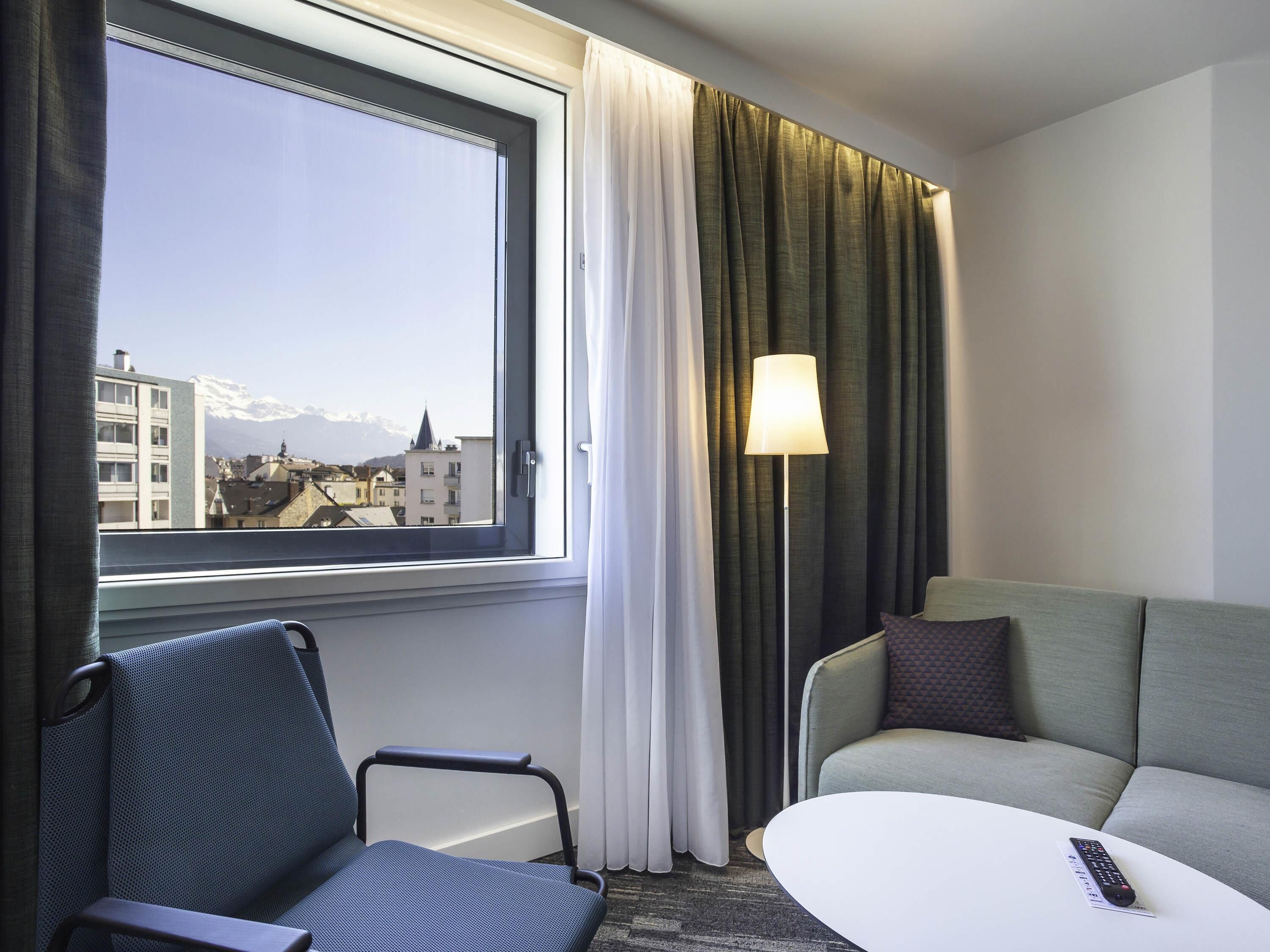 A hotel room with a large window, a sofa, a chair, a lamp, and a remote control on a small table.