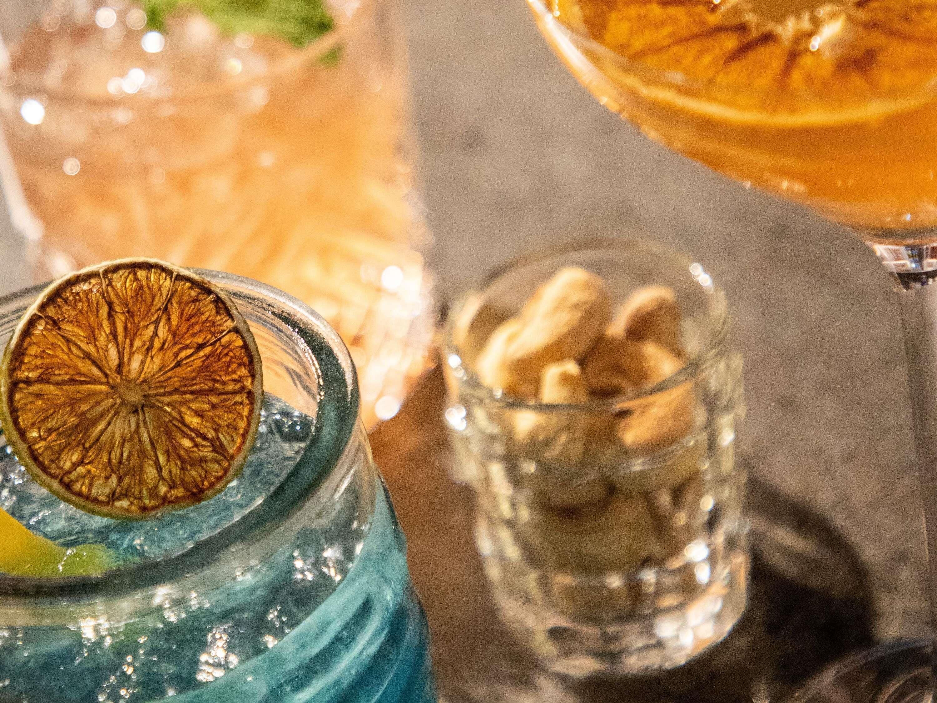 Close-up de um copo com uma fatia de laranja seca como guarnição, um copo com líquido azul e um copo pequeno com frutos secos.