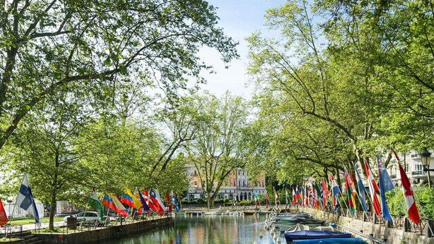A canal with boats docked on the right, surrounded by trees and flags. - Novotel Annecy Centre (Annecy)