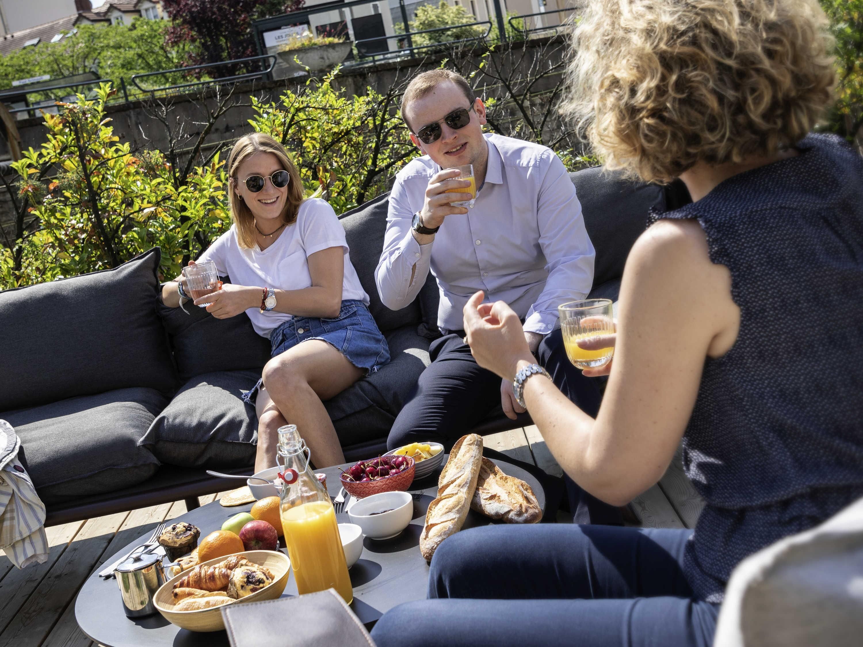 A group of people sitting on a grey outdoor sofa with a table in front of them, holding drinks and eating.