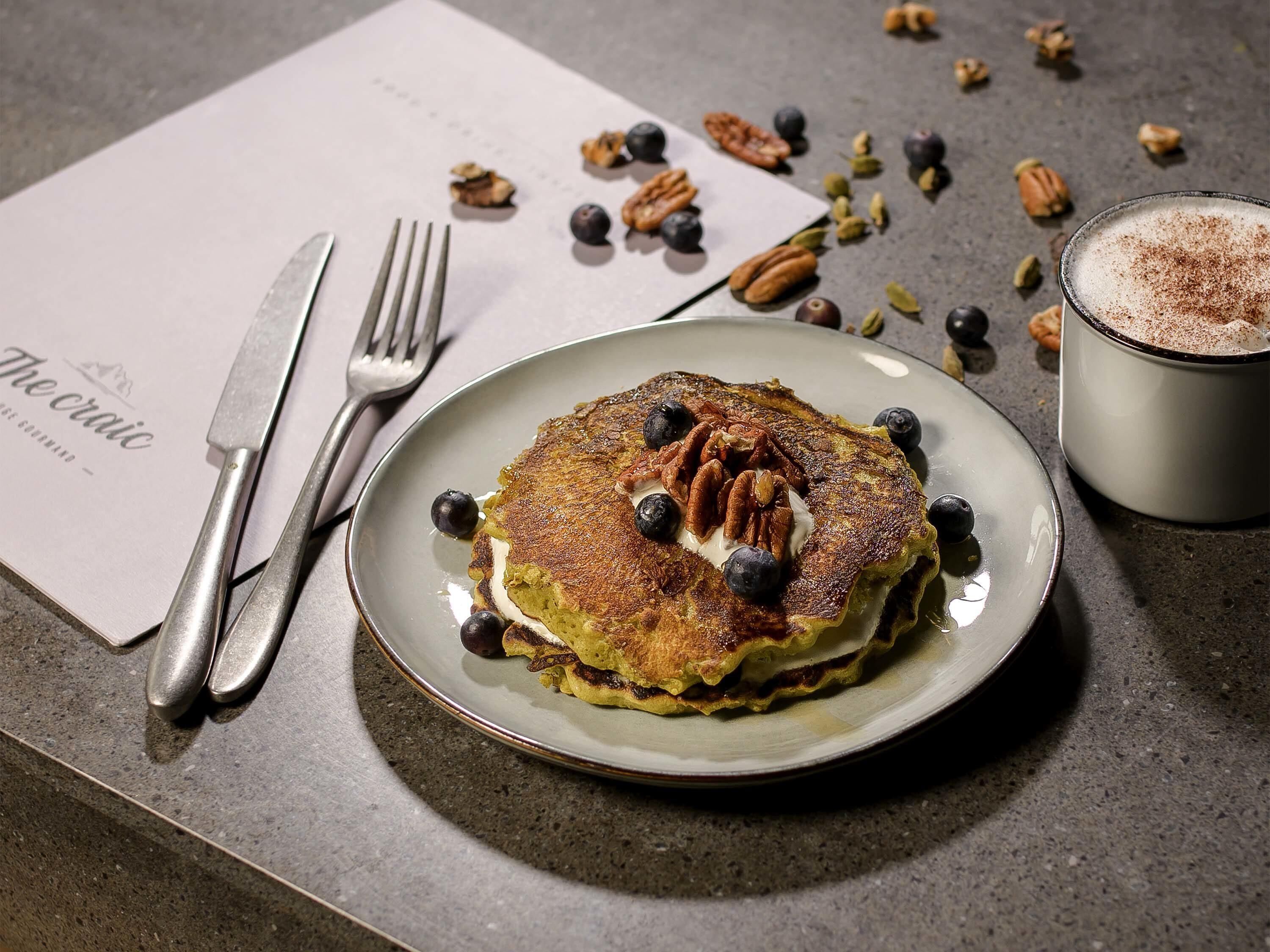 A plate of pancakes with blueberries and pecans, a fork, a knife, and a cup of coffee on a table.