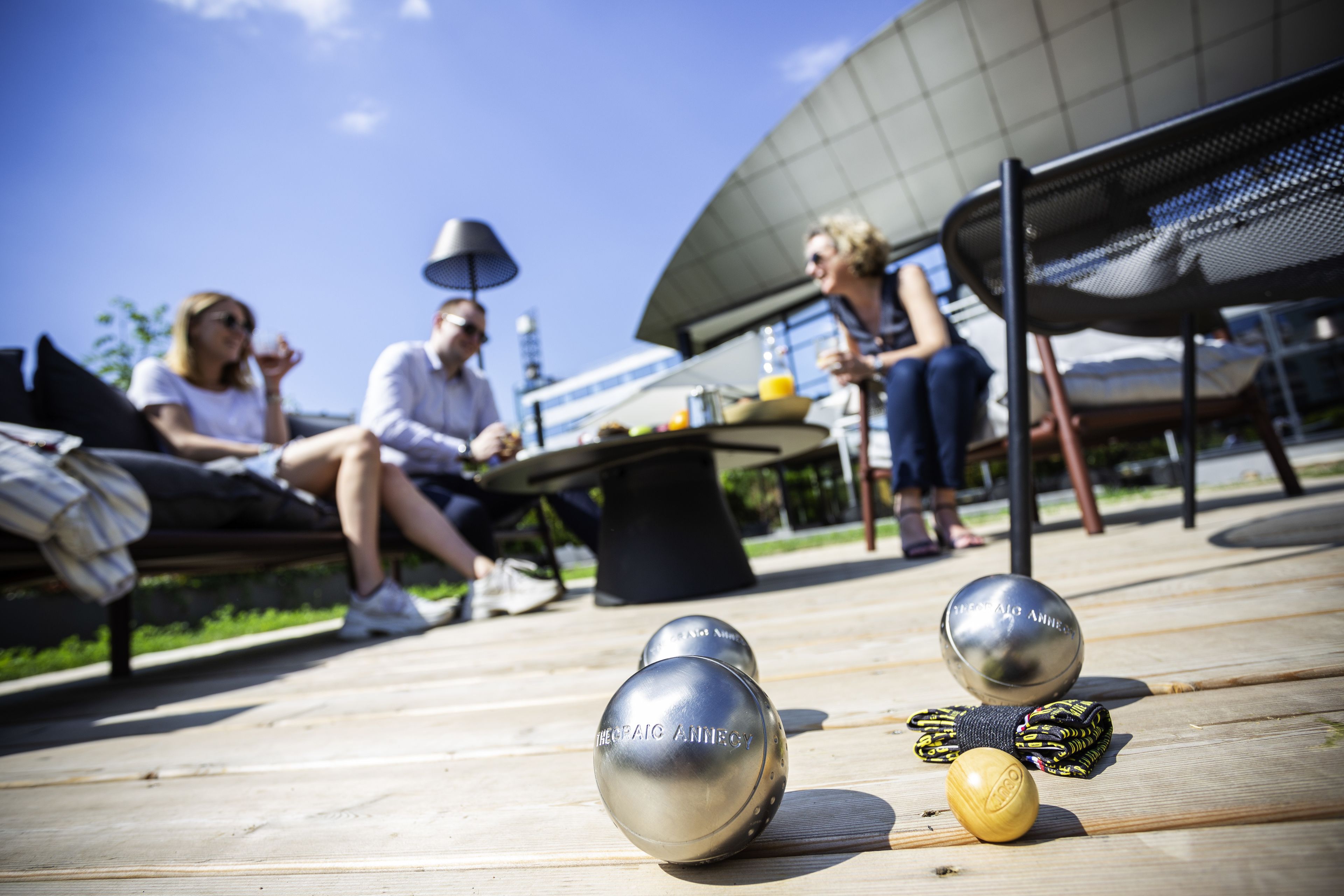 Un espacio al aire libre soleado con terraza de madera, sillas de metal y una mesa con lámpara.