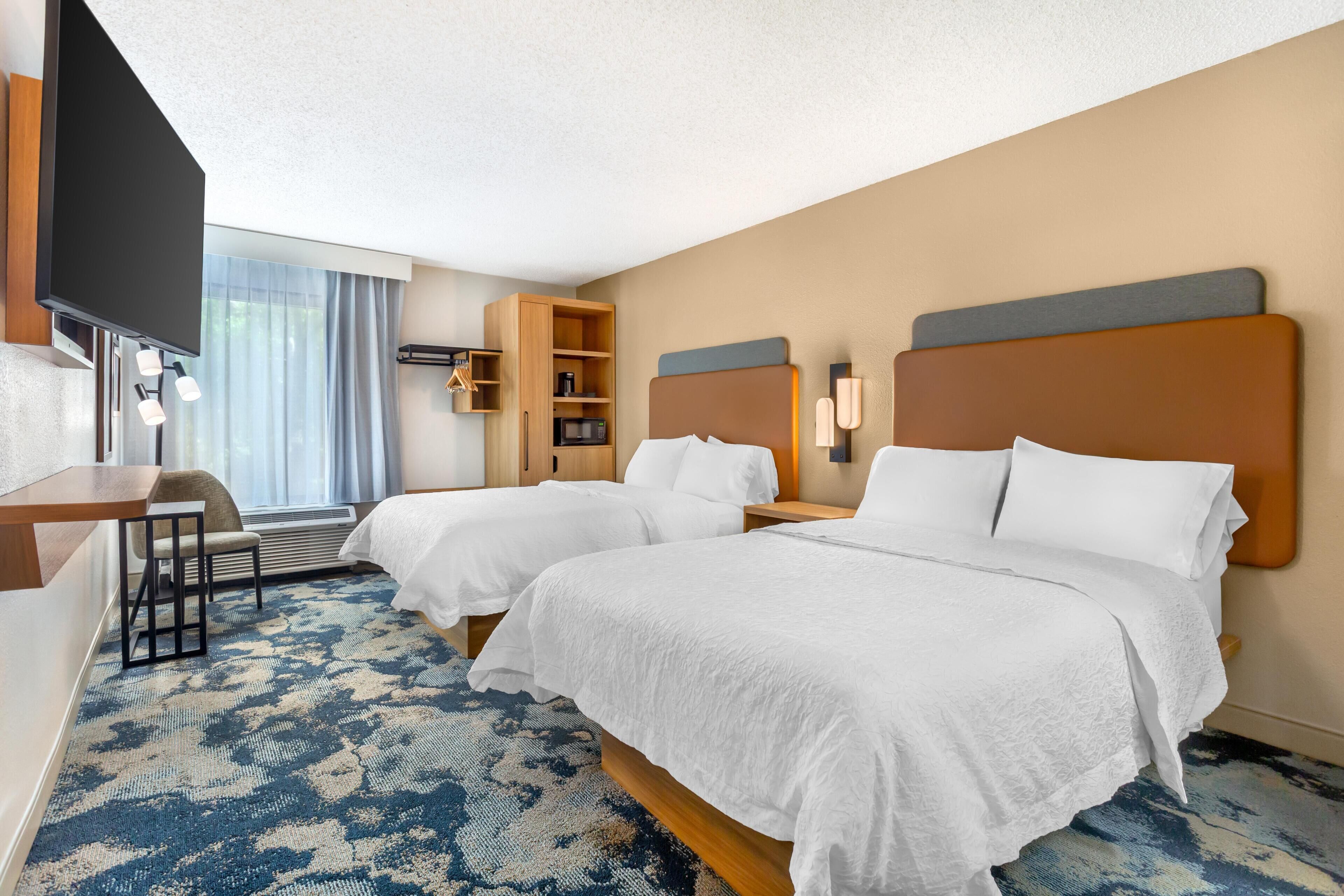 A hotel room with two beds, a desk, a chair, a TV, and a large window with curtains.