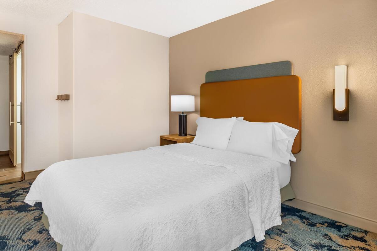 Hotelkamer met een groot bed, een nachtkastje met een lamp, een wandlamp en een deur naar een andere kamer.