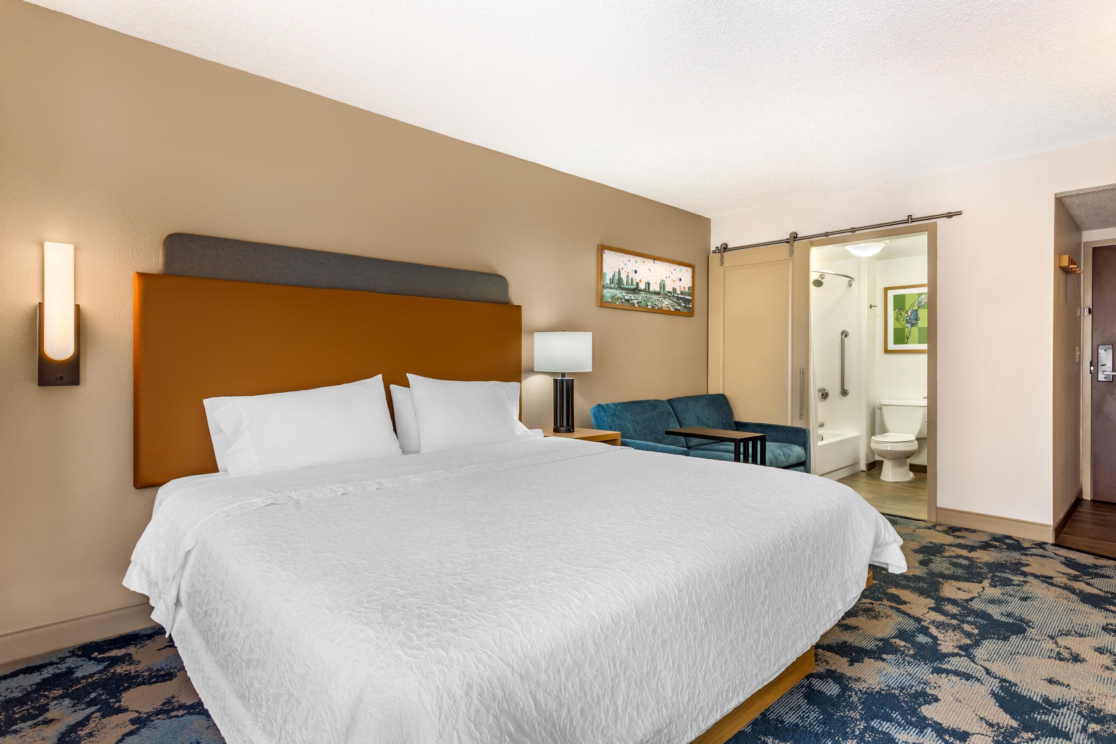 Habitación de hotel con una cama grande, una mesita de noche, una lámpara, una silla y un baño visible a través de una puerta abierta.
