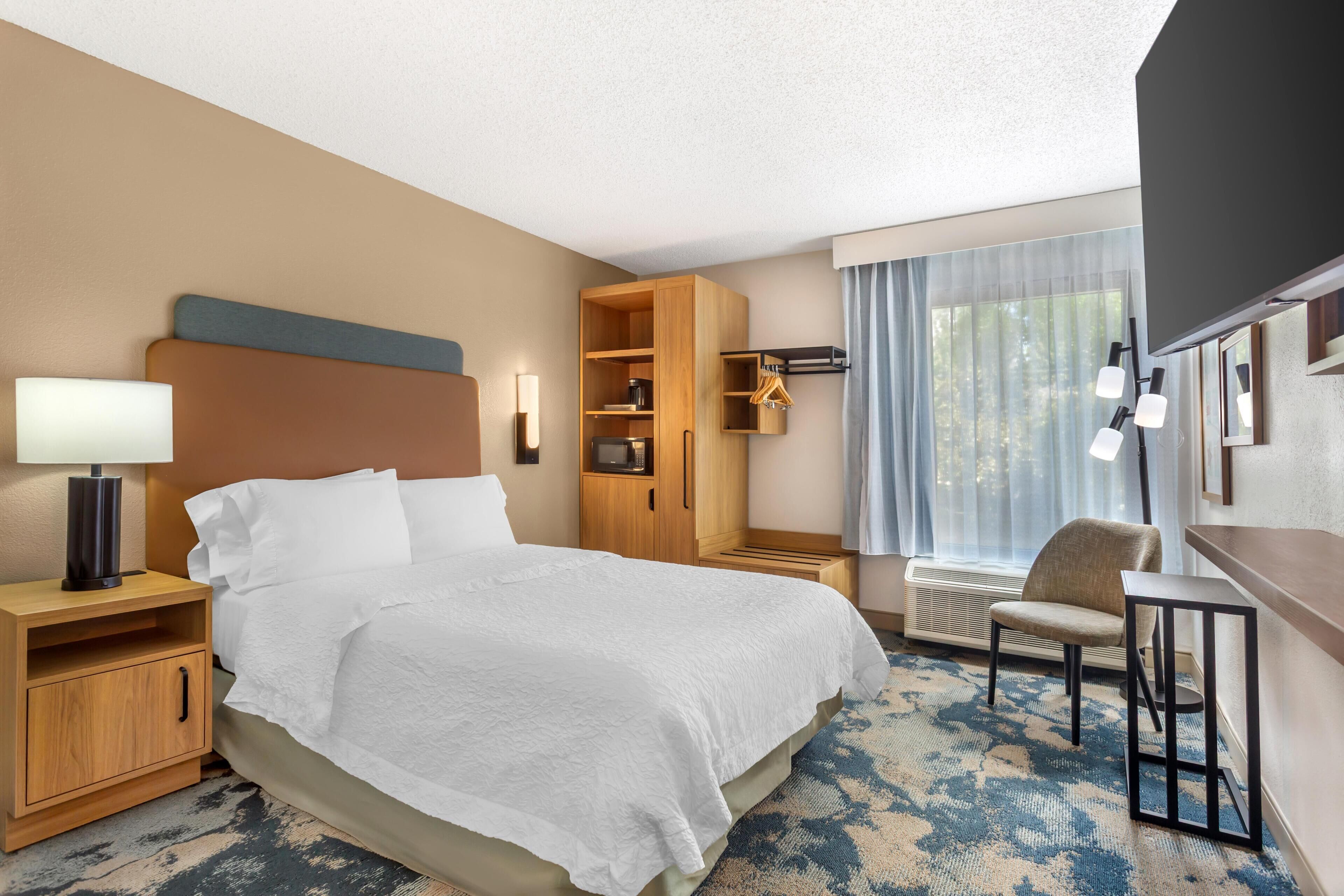 A hotel room with a bed, bedside table, lamp, wardrobe, chair, and a large window with curtains.