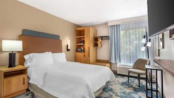 A hotel room with a bed, bedside table, lamp, wardrobe, chair, and a large window with curtains.