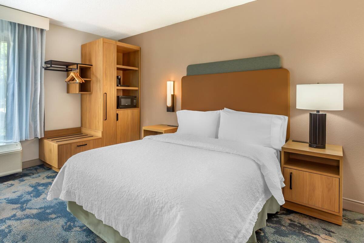 Een hotelkamer met een bed, een houten kledingkast, nachtkastje, lamp en een raam met gordijnen.