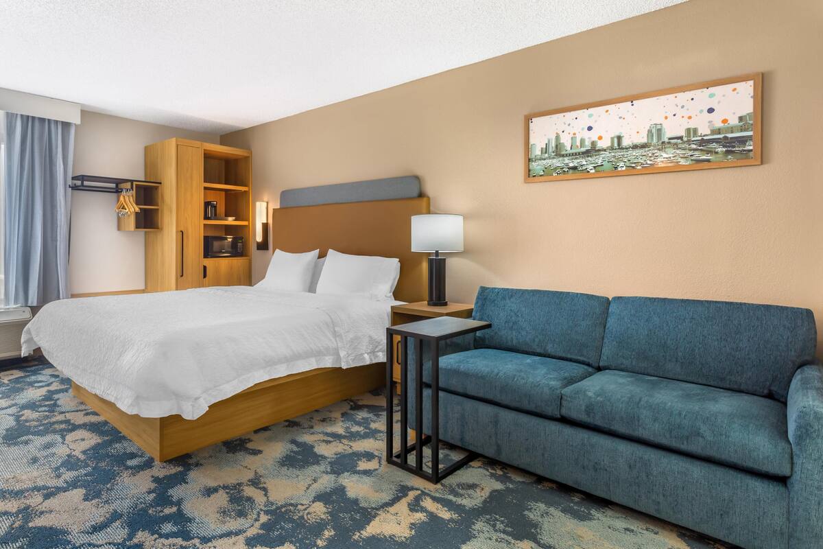 Een hotelkamer met een bed, een blauwe bank, een nachtkastje met lamp, een houten kast en een stadslandschap schilderij aan de muur.
