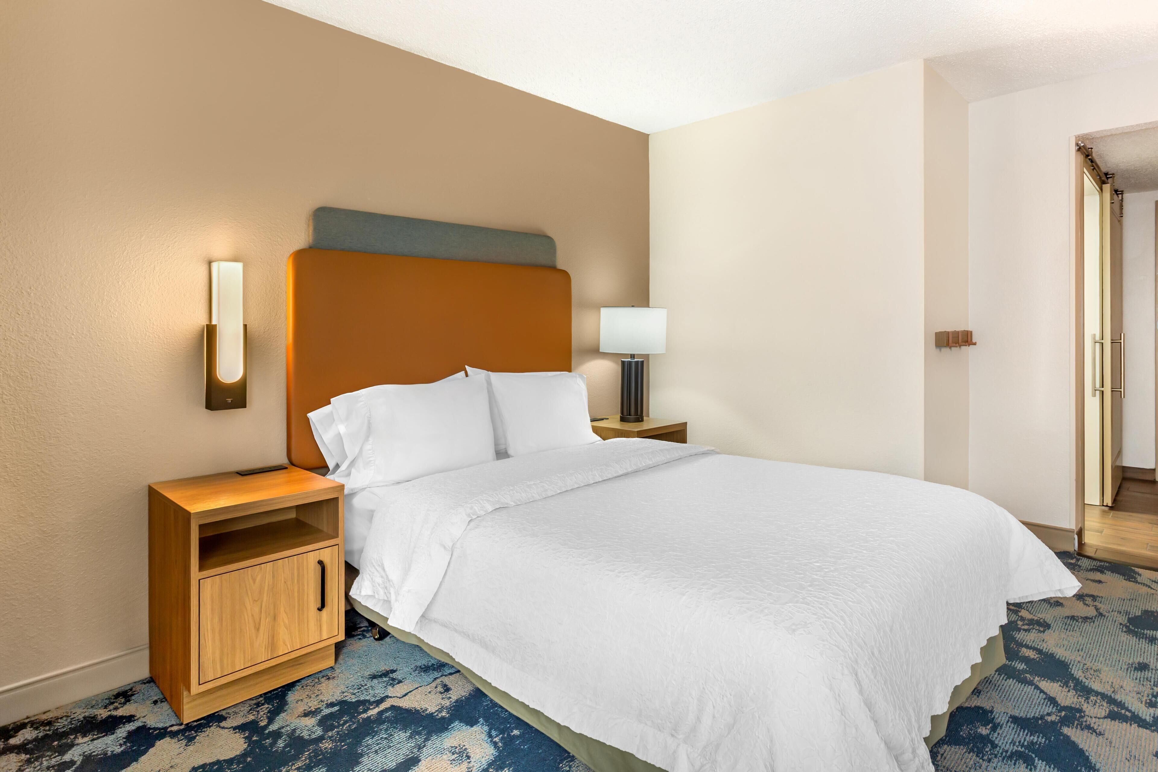 Habitación de hotel con una cama grande, una mesita de noche con lámpara, un armario de madera y una lámpara de pared.