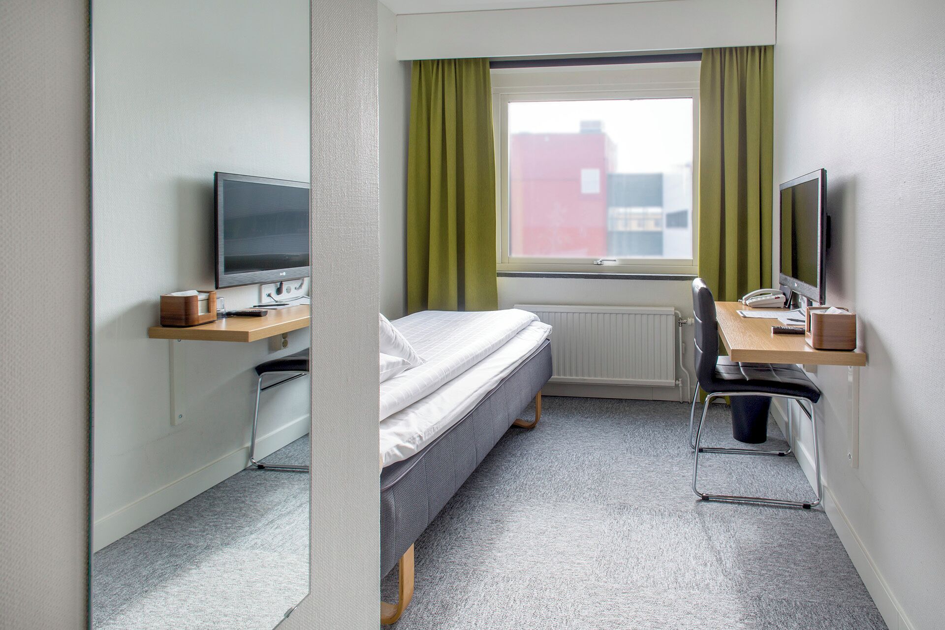 Une chambre d’hôtel de taille réduite, comprenant un lit, un bureau avec un ordinateur, une télévision et une fenêtre avec des rideaux.