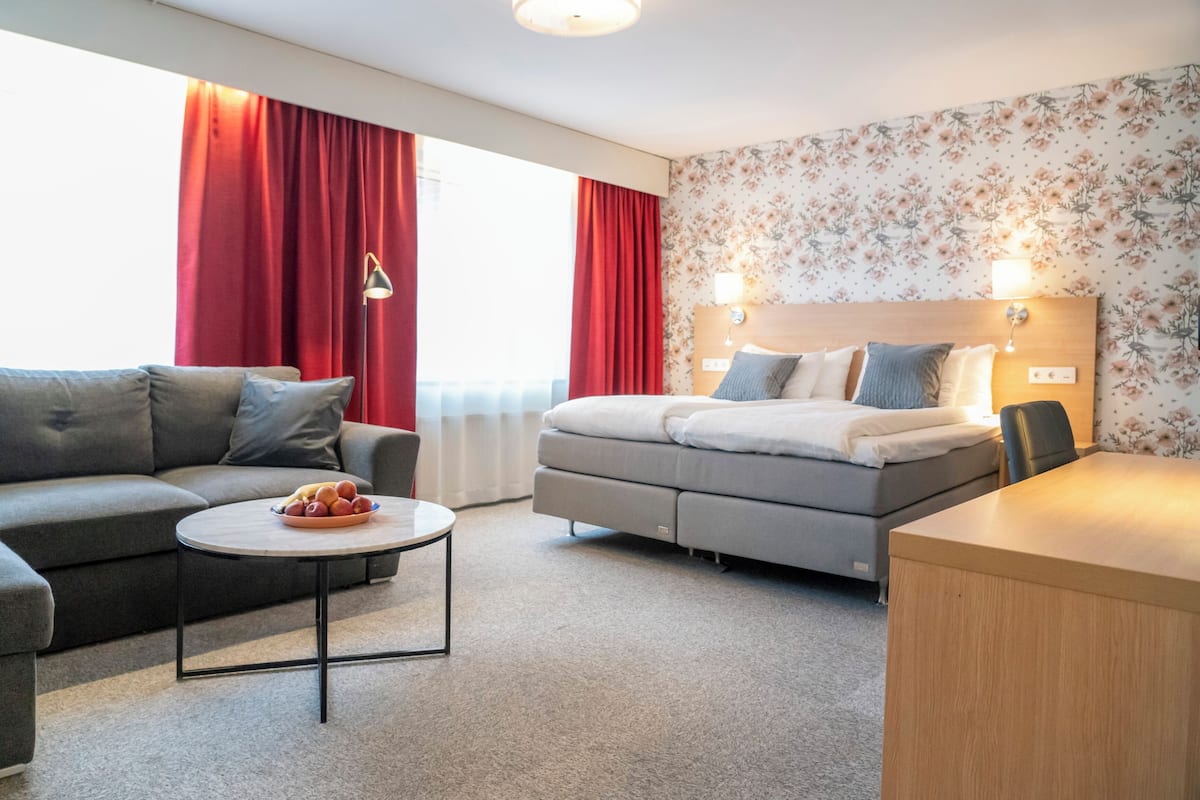 A modern hotel room with a grey sofa, a round coffee table with fruit, a bed with a wooden headboard, and a floral wallpaper.