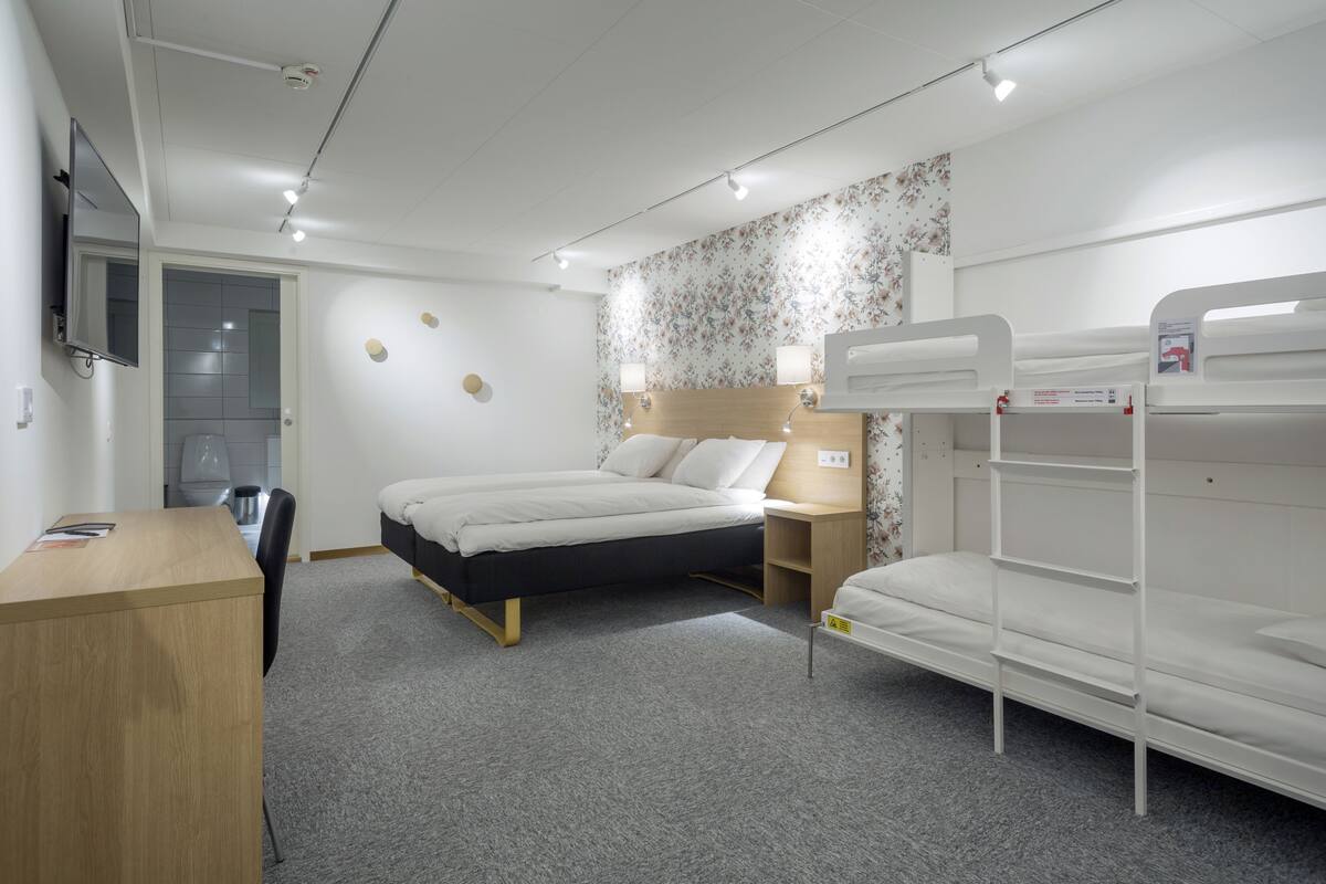 A hotel room with a bed, a desk, a chair, a TV, and a bunk bed.
