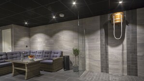 A modern bathroom with a glass shower enclosure, a wooden bucket showerhead, and a seating area with a sofa and table. - First Hotel Dragonen (Umea)