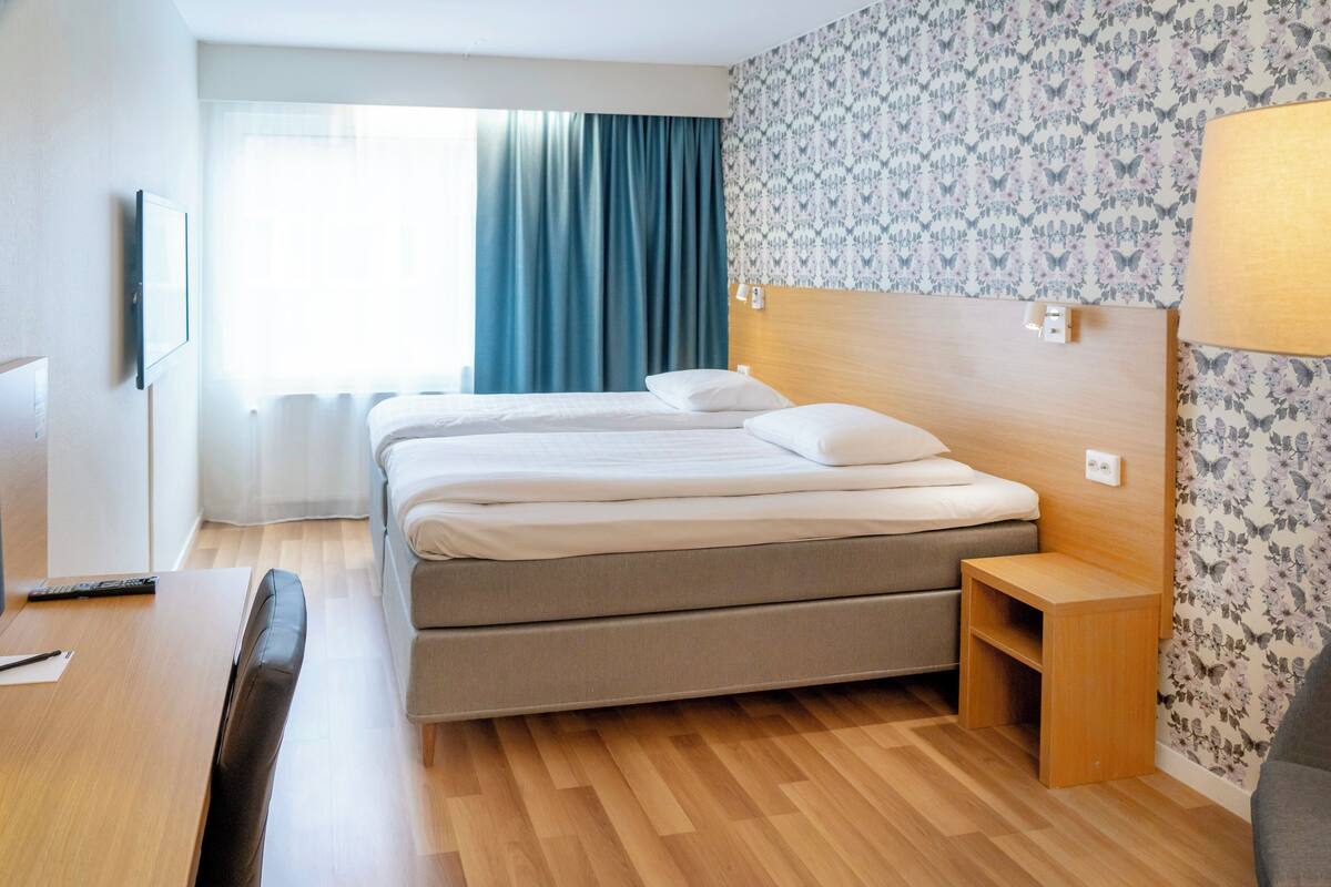 A hotel room with two beds, a wooden headboard, a desk with a chair, a lamp, and a window with curtains.