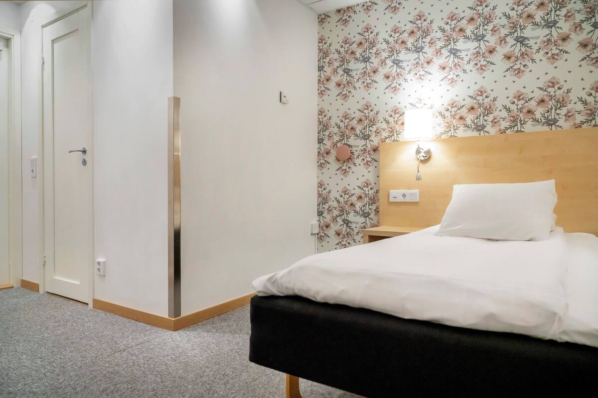 A hotel room with a bed, a nightstand, a wall-mounted light, and a floral wallpaper.