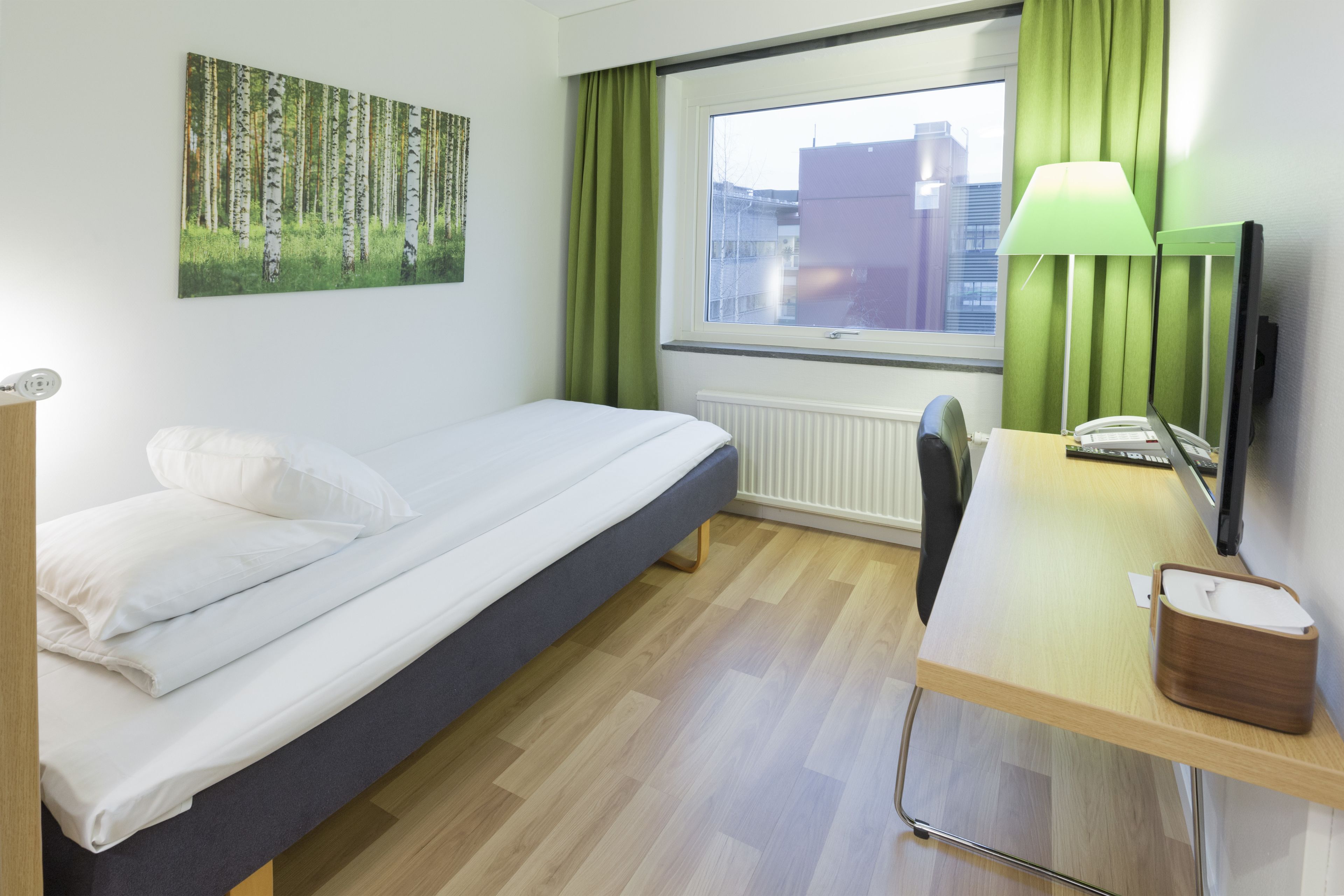 A hotel room with a bed, a desk with a lamp, a painting of a forest, and a window with curtains.
