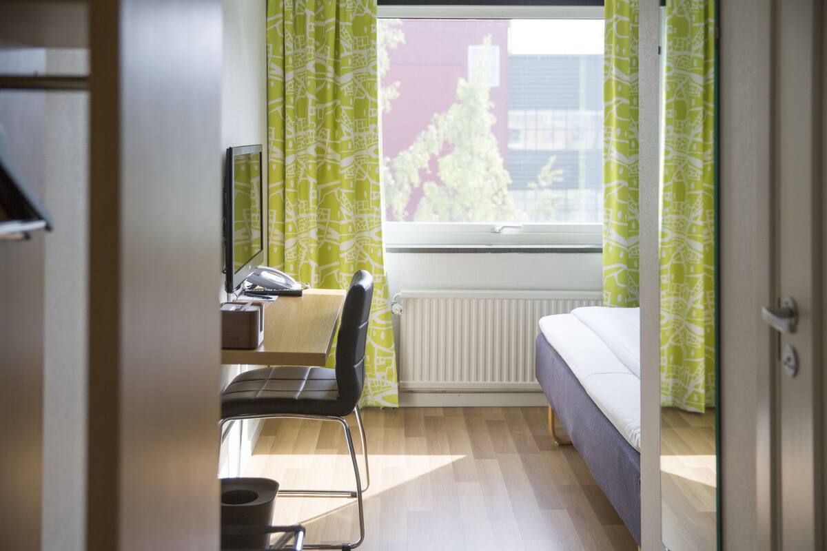 A bedroom with a bed, a desk, and a chair. There is a window with curtains, a radiator, and a view of trees and buildings outside.